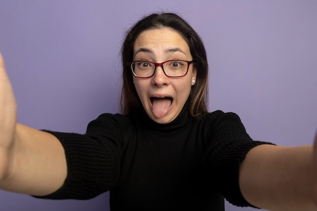 Free photo young beautiful girl in a black turtleneck and glasses happy and excited sticking out tongue