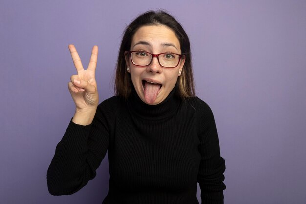 Young beautiful girl in a black turtleneck and glasses happy and positive sticking out tongue showing v-sign 