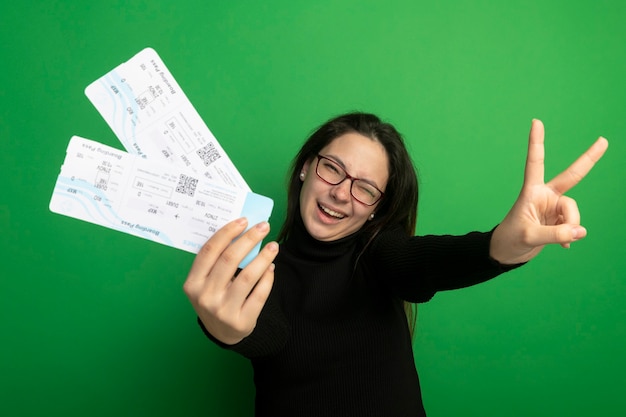 Young beautiful girl in a black turtleneck and glasses holding air tickets smiling with happy afce showing v-sign 