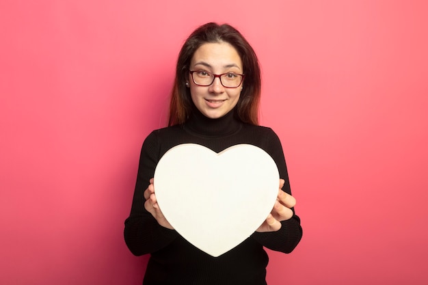 Free photo young beautiful girl in a black turtleneck and glasses holding heart shaped box smiling