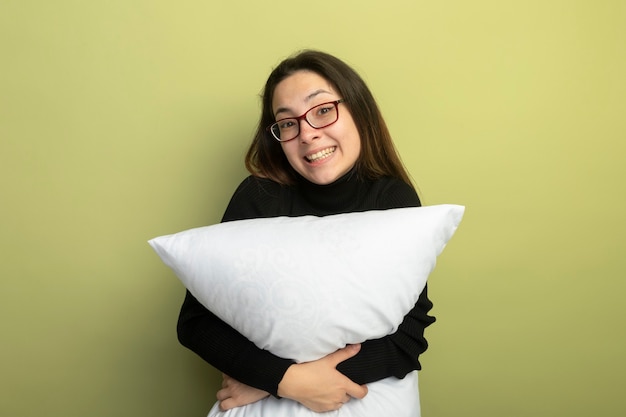 Free photo young beautiful girl in a black turtleneck and glasses holding pillow smilign with happy face