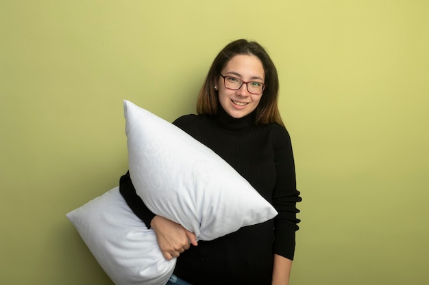 Young beautiful girl in a black turtleneck and glasses holding pillow smiling with happy face 