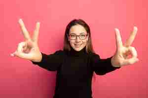 Free photo young beautiful girl in a black turtleneck and glasses lookign at camera smiling cheerfully showing v-sign