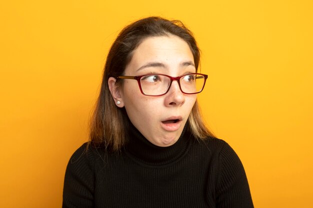 Young beautiful girl in a black turtleneck and glasses looking aside amazed and surprised 