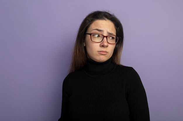 Young beautiful girl in a black turtleneck and glasses looking aside with skeptic expression