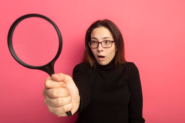 Young beautiful girl in a black turtleneck and glasses looking at something through magnifying glass being surprised 