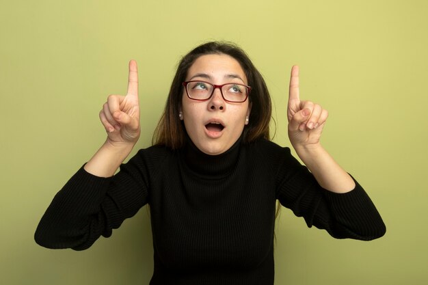 Young beautiful girl in a black turtleneck and glasses looking up pointgn with index figners up 