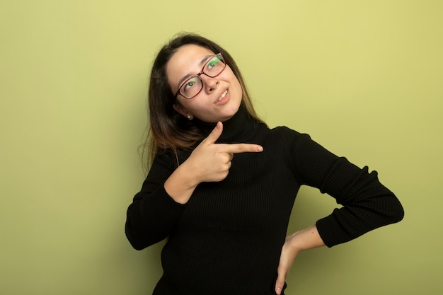 Young beautiful girl in a black turtleneck and glasses looking up puzzled pointing with index figner to the side 