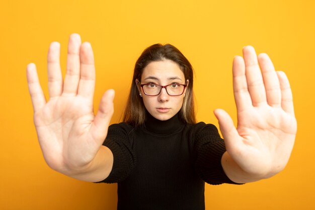 Young beautiful girl in a black turtleneck and glasses making stop sing with hands with fear expression 