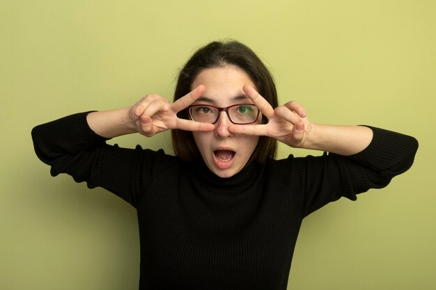 Young beautiful girl in a black turtleneck and glasses making v-sign with both hands looking through fingers 