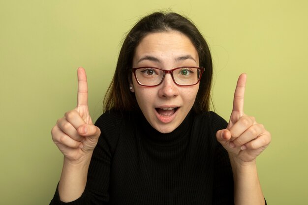 Young beautiful girl in a black turtleneck and glasses pointing with index fingers up smiling 