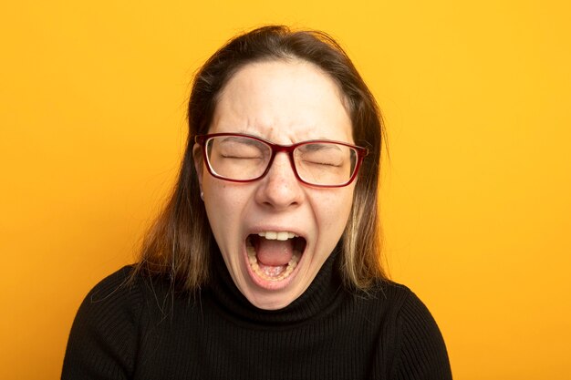 Young beautiful girl in a black turtleneck and glasses shouting with annoyed expression going wild 