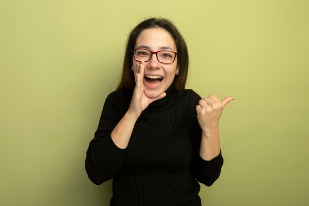 Young beautiful girl in a black turtleneck and glasses shouting with hand near mouth pointing with finger to the side 