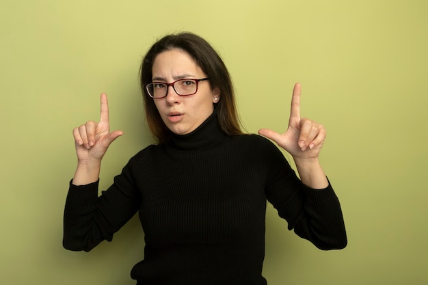 Young beautiful girl in a black turtleneck and glasses showing index fingers with serious expression