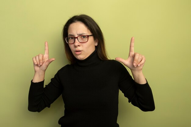 Young beautiful girl in a black turtleneck and glasses showing index fingers with serious expression
