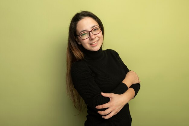 Young beautiful girl in a black turtleneck and glasses smiling confident with crossed arms on chest 