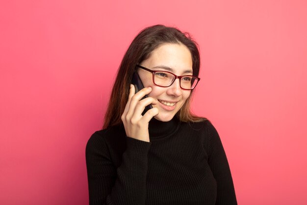 Young beautiful girl in a black turtleneck and glasses talking on mobile phone smiling with happy face 