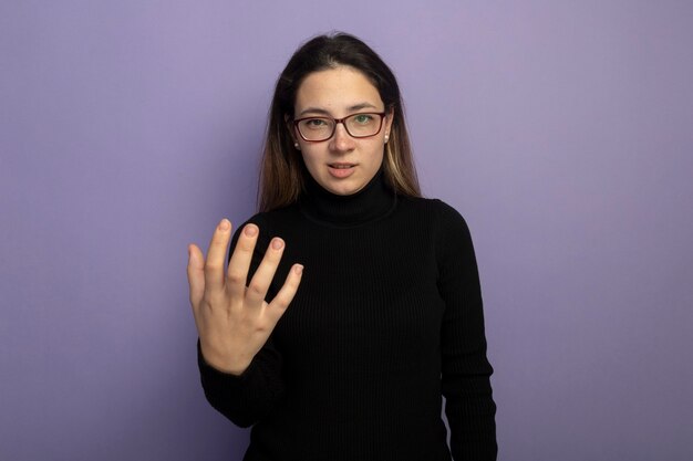 Young beautiful girl in a black turtleneck and glasses with arm out being displeased 