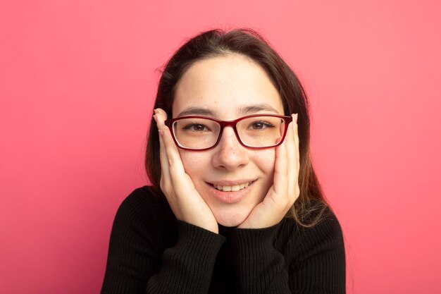 Young beautiful girl in a black turtleneck and glasses with hands on face smiling cheerfully 