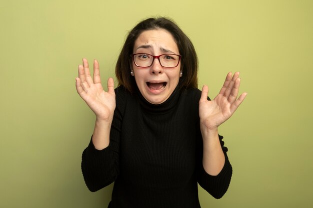 Young beautiful girl in a black turtleneck and glasses yelling with raised hands being disappointed 