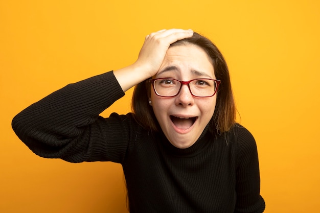 Young beautiful girl in a black turtleneck lookign at camera confused shouting with hand on her head for mistake 