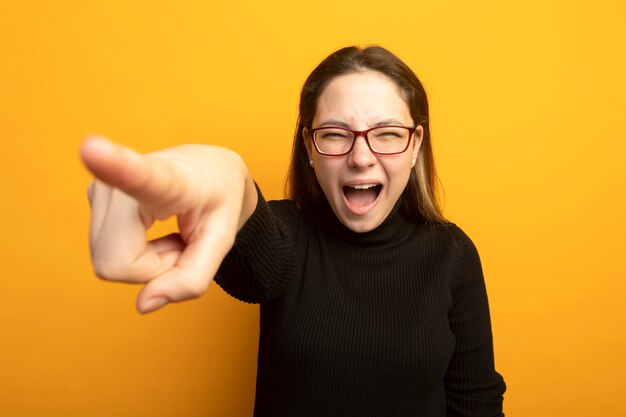 Young beautiful girl in a black turtleneck pointing with index figner at camera shouting 