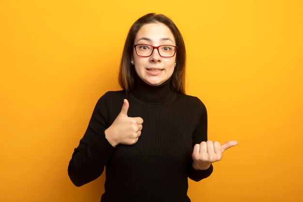 Young beautiful girl in a black turtleneck pointing with thumb to the side showing thumbs up 