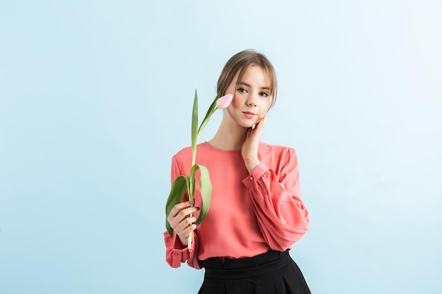 Young beautiful girl in blouse holding pink tulip in hand near face while dreamily looking in camera over blue background