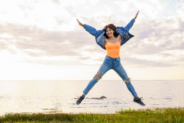 Free photo a young beautiful girl in a denim jacket, jeans and a yellow t-shirt jumps on the background of the sea on a summer day, posing at sunset