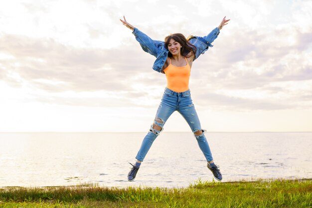 A young beautiful girl in a denim jacket, jeans and a yellow t-shirt jumps over the sea on a summer day, posing at sunset