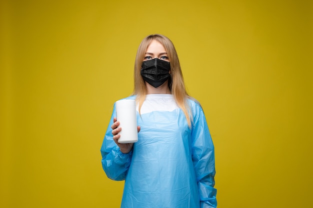 Young beautiful girl in a disposable medical gown and with a mask on her face holds wet antibacterial wipes, portrait isolated on yellow background