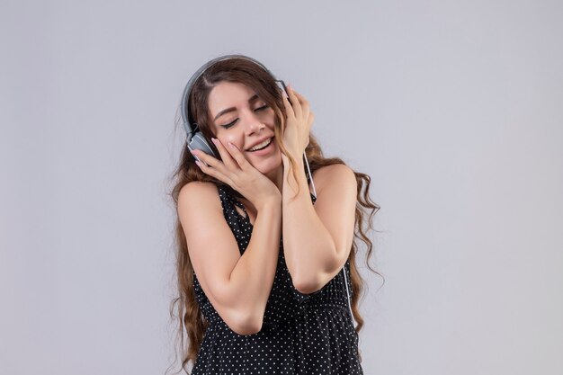 Young beautiful girl in dress in polka dot enjoying favorite music through wireless headphones standing with closed eyes over white background