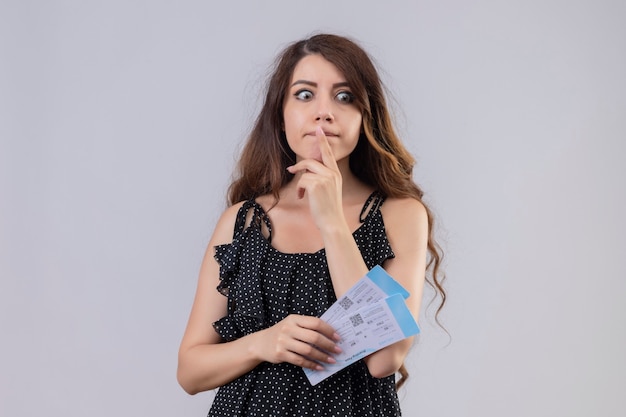 Young beautiful girl in dress in polka dot holding air tickets standing with pensive expression with finger on lips  thinking having doubts over white background