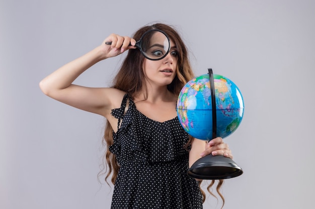 Young beautiful girl in dress in polka dot looking at globe through magnifying glass intrigued standing over white background