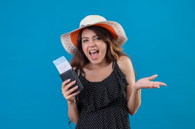 Young beautiful girl in dress in polka dot in summer hat holding air tickets exited and happy standing over blue background