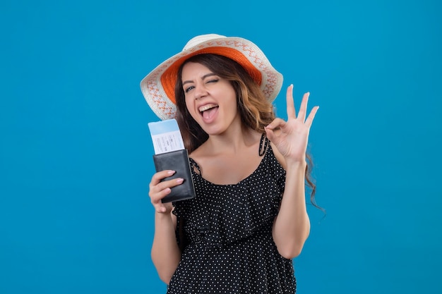 Young beautiful girl in dress in polka dot in summer hat holding air tickets exited and happy winking doing ok sign standing over blue background