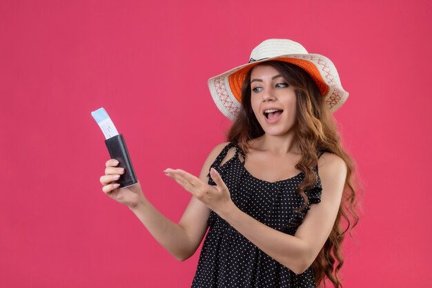 Young beautiful girl in dress in polka dot in summer hat holding air tickets presenting with arm of her hand smiling cheerfully positive and happy standing over pink background