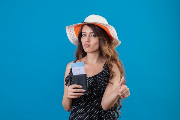 Free photo young beautiful girl in dress in polka dot in summer hat holding air tickets showing thumbs up positive and happy standing over blue background