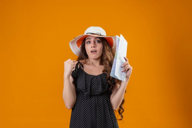 Young beautiful girl in dress in polka dot in summer hat holding map raising fist confused with skeptic expression on face standing over yellow background