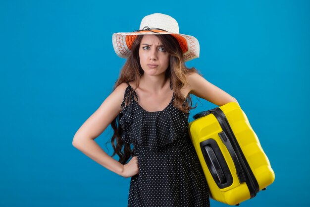 Young beautiful girl in dress in polka dot in summer hat holding suitcase looking at camera with angry face standing over blue background