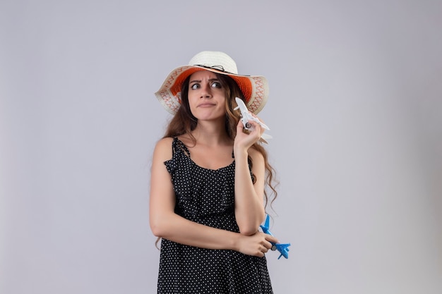 Young beautiful girl in dress in polka dot in summer hat holding toy airplanes looking aside displeased with skeptic expression on face standing over white background