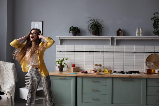 Foto gratuita giovane bella ragazza in abiti alla moda che si diverte in cucina modello bruna felice con i capelli lunghi che ascolta musica e balla donna alla moda in giacca gialla e cuffie blu che cantano