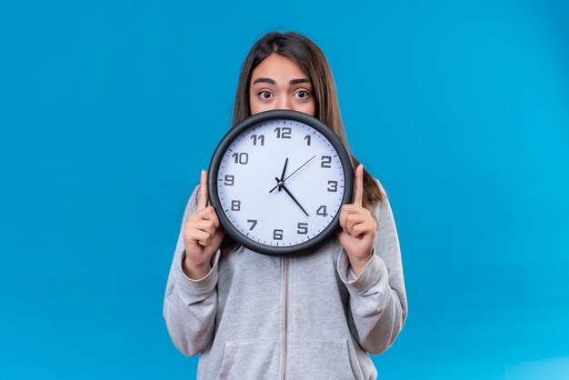 Young beautiful girl in gray hoody holding clock and looking at camera with surprised sight standing over blue background