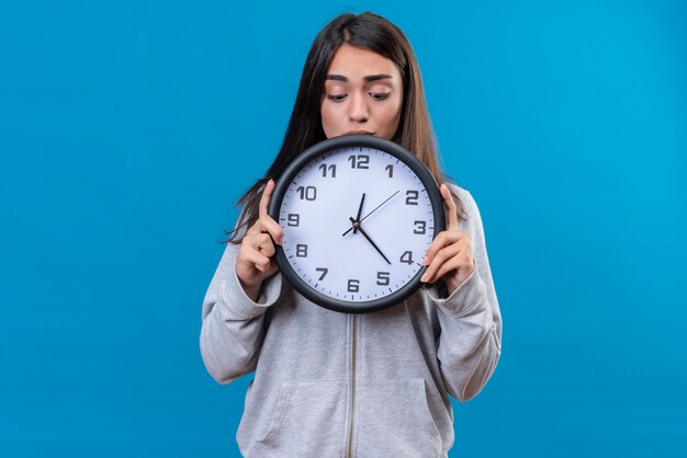 Young beautiful girl in gray hoody holding clock and looking with confusion sight to clock standing over blue background