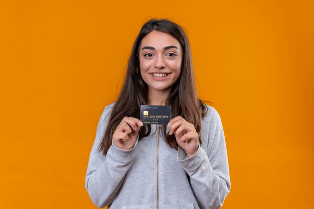 Young beautiful girl in gray hoody holding credit card and looking at camera with simle on face standing over orange background