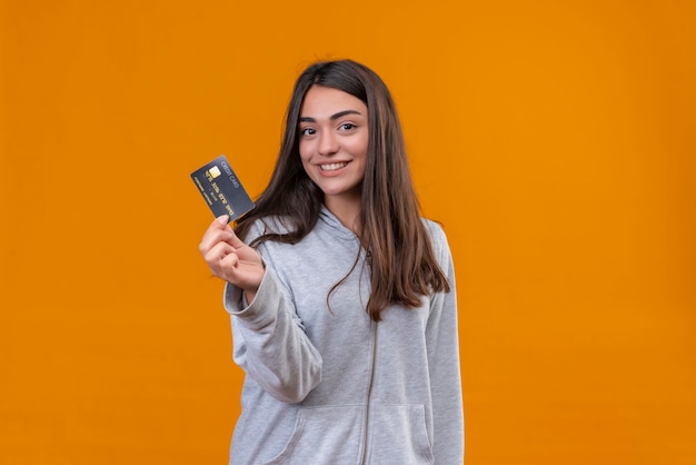 Young beautiful girl in gray hoody holding credit card and looking at camera with smile standing over orange background