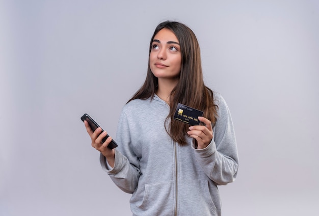 Молодая красивая девушка в серой толстовке с капюшоном держит телефон и кредитную карту, глядя в сторону с задумчивым выражением лица, стоя на белом фоне