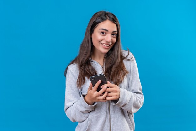Young beautiful girl in gray hoody holding telephone looking at camera with pleasure standing over blue background