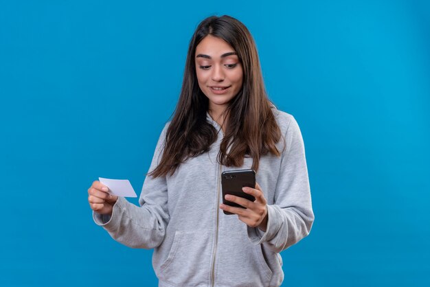 Молодая красивая девушка в серой толстовке с капюшоном держит телефон и бумагу, глядя на телефонный сюрприз на лице, стоящем на синем фоне