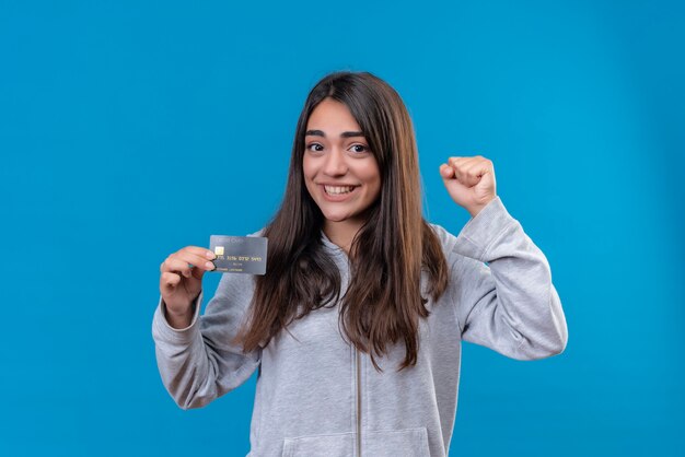 Young beautiful girl in gray hoody holding telephone rejoicing her success and victory exited and happy standing over blue background